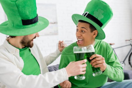 Excited interracial friends clinking green beer during saint patrick day at home 
