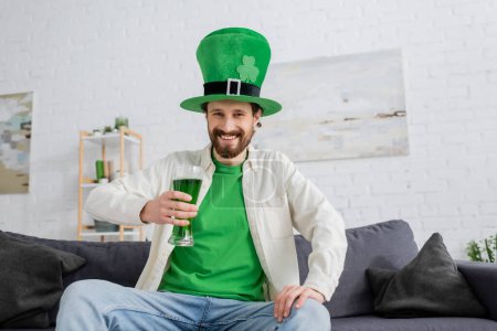 Hombre sonriente con sombrero verde sosteniendo cerveza y mirando a la cámara durante la celebración de San Patricio en casa 