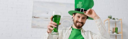 Hombre positivo sosteniendo cerveza verde y sombrero durante el día de San Patricio en casa, estandarte 