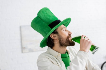 Foto de Vista lateral del hombre en sombrero bebiendo cerveza verde durante la celebración de San Patricio en casa - Imagen libre de derechos