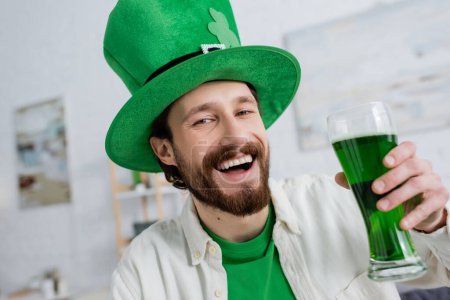 Joyeux homme en chapeau saint Patrick tenant de la bière verte et regardant la caméra à la maison 