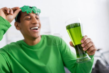 Verschwommener Afroamerikaner mit Sonnenbrille und grünem Bier zu Hause 