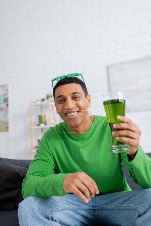 Cheerful african american man holding green beer and looking at camera at home 