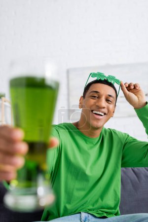 Positve african american man holding party sunglasses and blurred beer at home 