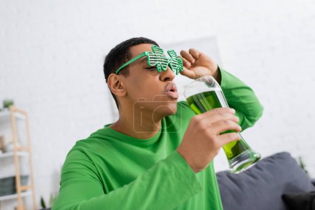 Hombre afroamericano en gafas de sol de fiesta sosteniendo cerveza verde durante el día de San Patricio