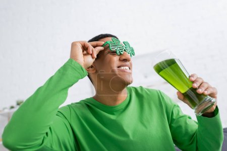 Souriant homme afro-américain dans des lunettes de soleil de fête tenant la bière verte pendant la Saint Patrick Day