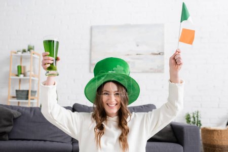 Fröhliche junge Frau mit grünem Hut mit irischer Flagge und Bier zu Hause 