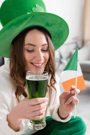 Positive woman holding green beer and Irish flag while celebrating saint patrick day