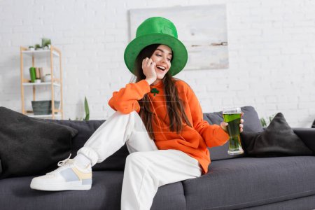 Positive woman in green hat holding glass of green beer on couch at home 