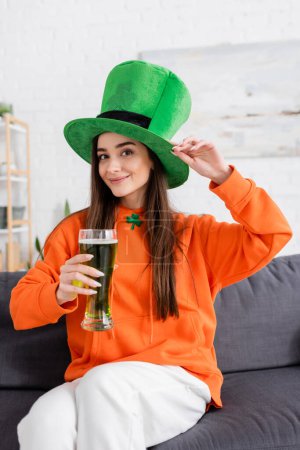 Mujer joven positiva con sombrero verde sosteniendo cerveza mientras está sentada en el sofá 