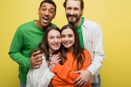 Cheerful interracial men hugging friends isolated on yellow 