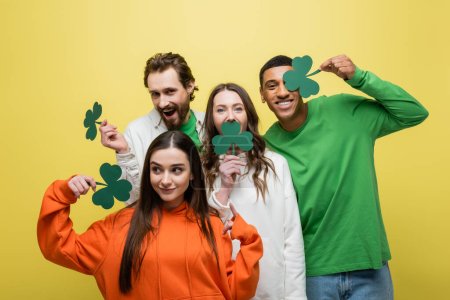 Multiethnic friends holding paper clover during saint patrick day isolated on yellow 