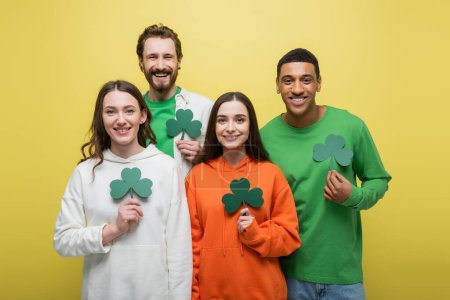Happy multiethnic people holding paper clover isolated on yellow 