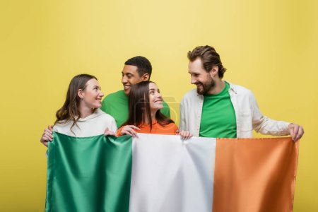 Interracial friends looking at each other while holding Irish flag isolated on yellow 