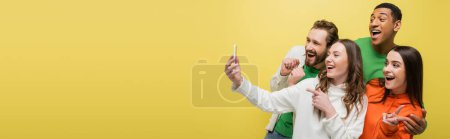 Multiethnic friends using cellphone and gesturing isolated on yellow, banner 