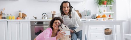 African american couple taking selfie with labrador in kitchen at home, banner 