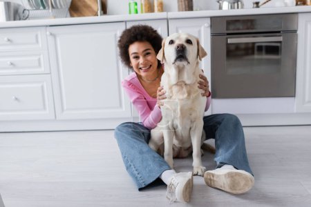 Young african american woman petting labrador on floor in kitchen 