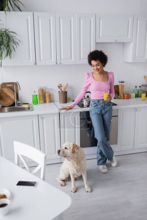 Positive african american woman holding orange juice near labrador in kitchen 