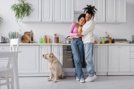 Cheerful african american couple hugging near labrador in kitchen 