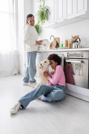 Cheerful african american woman petting labrador near boyfriend washing plate in kitchen 