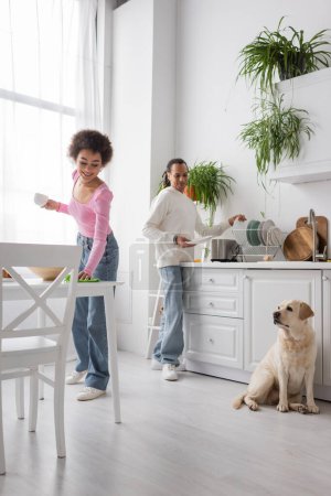 Positive african american woman holding cup and cleaning table near boyfriend and labrador in kitchen 