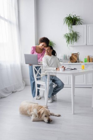 Smiling african american woman holding laptop near boyfriend and labrador at home  