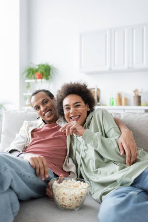 Cheerful african american woman holding popcorn and looking at camera near boyfriend on couch 