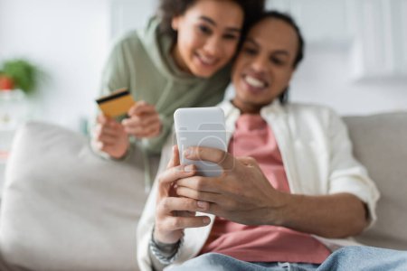 Foto de Pareja afroamericana borrosa usando teléfono inteligente y tarjeta de crédito durante las compras en línea en casa - Imagen libre de derechos