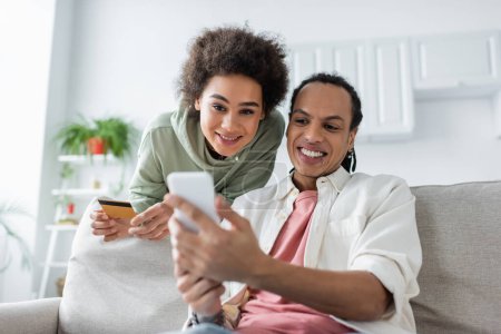 Téléchargez les photos : Sourire couple afro-américain en utilisant un smartphone lors des achats en ligne à la maison - en image libre de droit