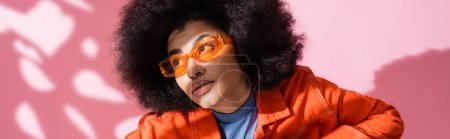 mujer americana africana rizada en gafas de sol naranjas elegantes mirando hacia otro lado en rosa, bandera  
