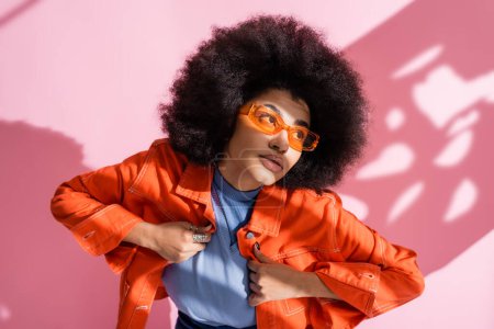 Photo for Curly african american woman in stylish orange sunglasses and trendy outfit posing on pink - Royalty Free Image