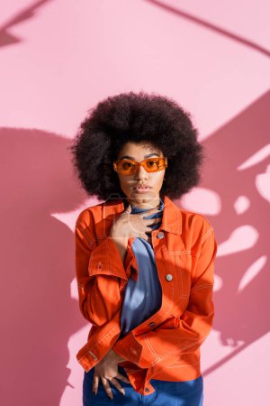 Foto de Mujer americana africana rizada en gafas de sol naranjas elegantes y traje de moda mirando a la cámara en rosa - Imagen libre de derechos