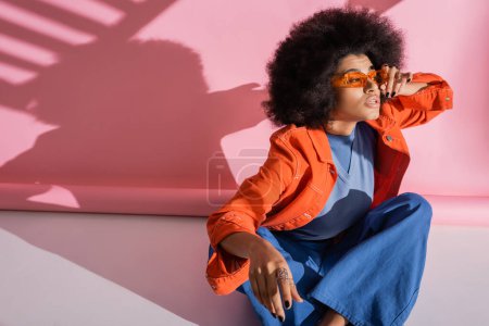 Foto de Mujer afroamericana rizada en traje de moda ajustando gafas de sol naranjas en rosa - Imagen libre de derechos