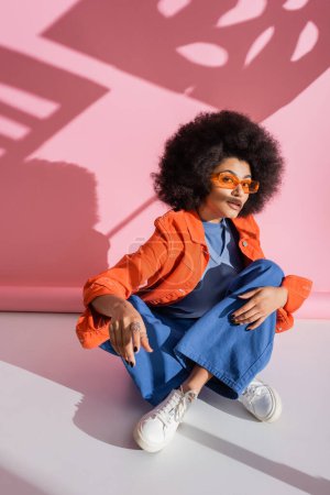 Foto de Mujer americana africana rizada en gafas de sol naranjas elegantes y traje de moda sentado en rosa - Imagen libre de derechos