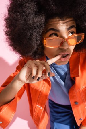 lockige afrikanisch-amerikanische Frau in stylischer orangefarbener Sonnenbrille beißt Finger und schaut weg auf rosa 