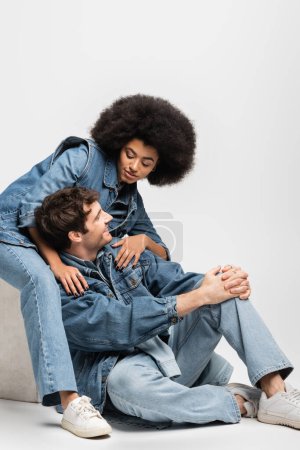 brunette african american woman in sneakers sitting and looking at cheerful man in denim outfit on grey 