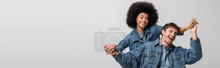 cheerful african american model looking at camera and holding hands with stylish man isolated on grey, banner 