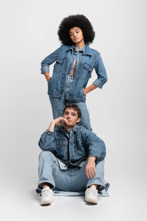 Téléchargez les photos : Full length of curly african american woman standing with hands in pockets behind man in denim outfit on grey - en image libre de droit