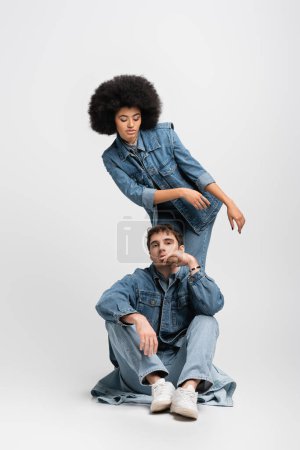 Téléchargez les photos : Full length of curly african american woman standing behind man in denim outfit and sneakers on grey - en image libre de droit