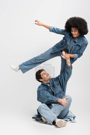 Foto de Full length of happy african american woman posing near cheerful man in denim outfit and sneakers sitting on grey - Imagen libre de derechos