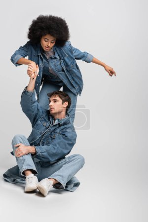 Foto de Full length of man in denim clothes sitting and holding hand of curly african american woman on grey - Imagen libre de derechos