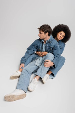 Foto de Full length of curly african american woman sitting and hugging stylish man in denim clothes and sneakers on grey - Imagen libre de derechos
