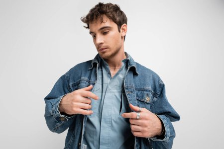 stylish young man adjusting denim jacket and looking away isolated on grey 