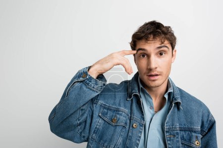 Foto de Overwhelmed man in denim jacket pointing with finger at head and looking at camera isolated on grey - Imagen libre de derechos