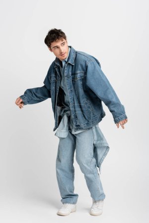 full length of stylish young man in denim jacket and jeans posing while looking away on grey 