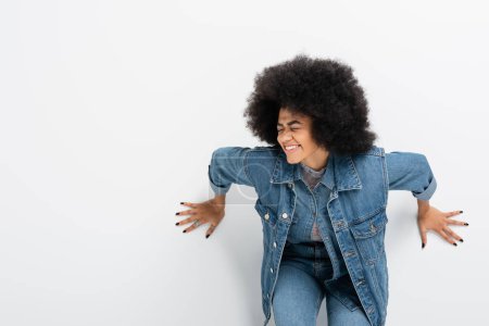 lockige Afroamerikanerin in Jeansjacke lächelt mit geschlossenen Augen gegen graue Wand 
