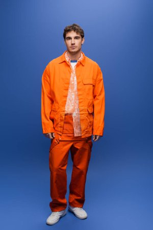 Full length of stylish man in orange jacket and overall standing on blue background