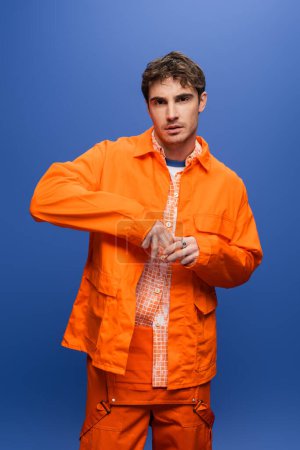 Trendy man posing in orange overall and jacket isolated on blue 
