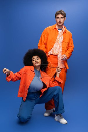 Stylish african american woman in jacket posing near man in overall on blue background