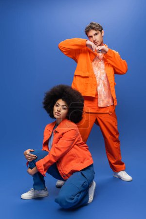 Trendy man in overalls posing near african american woman in orange jacket on blue background